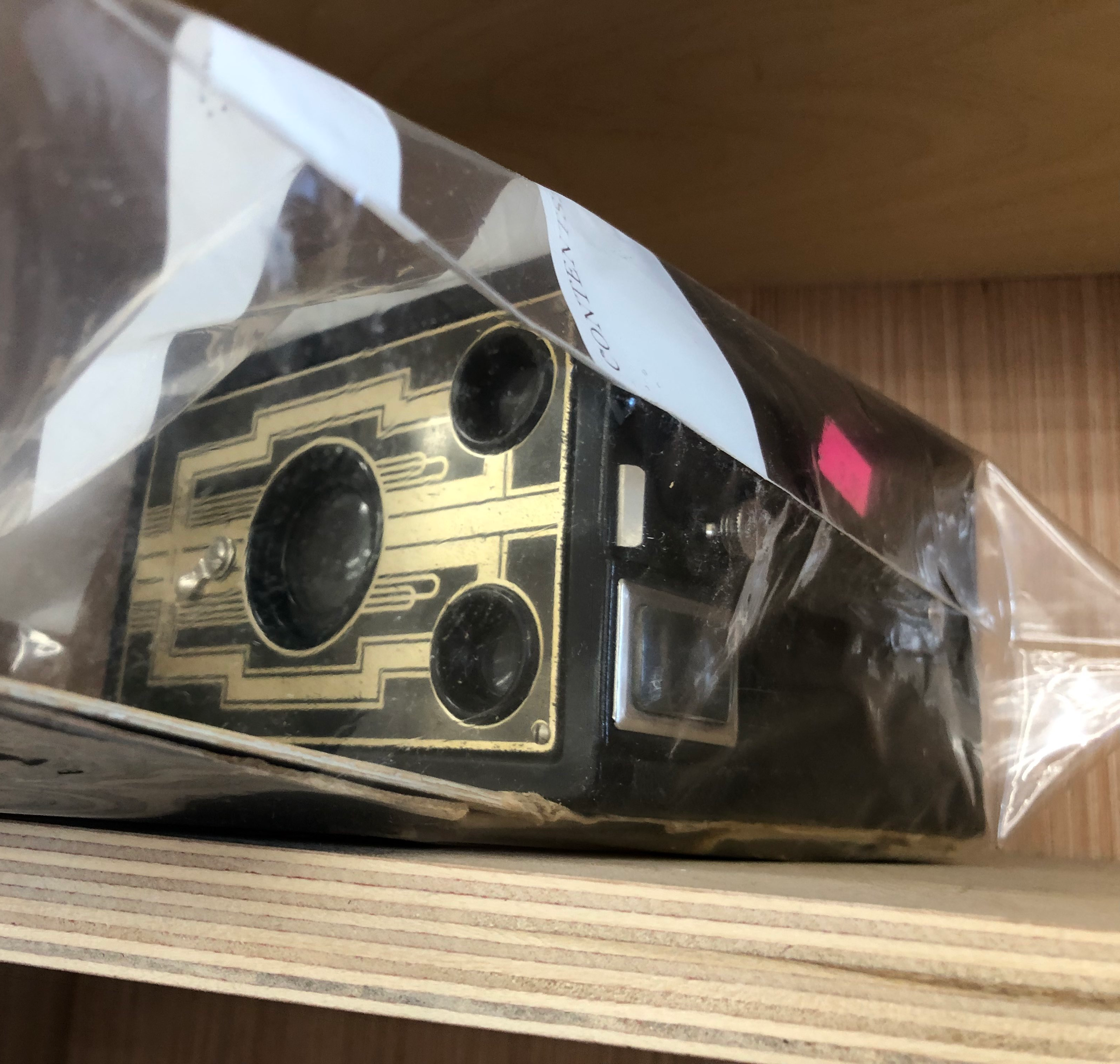 a black box camera with a geometric art deco design on the front. it is in a plastic bag ans sitting on a wooden shelf